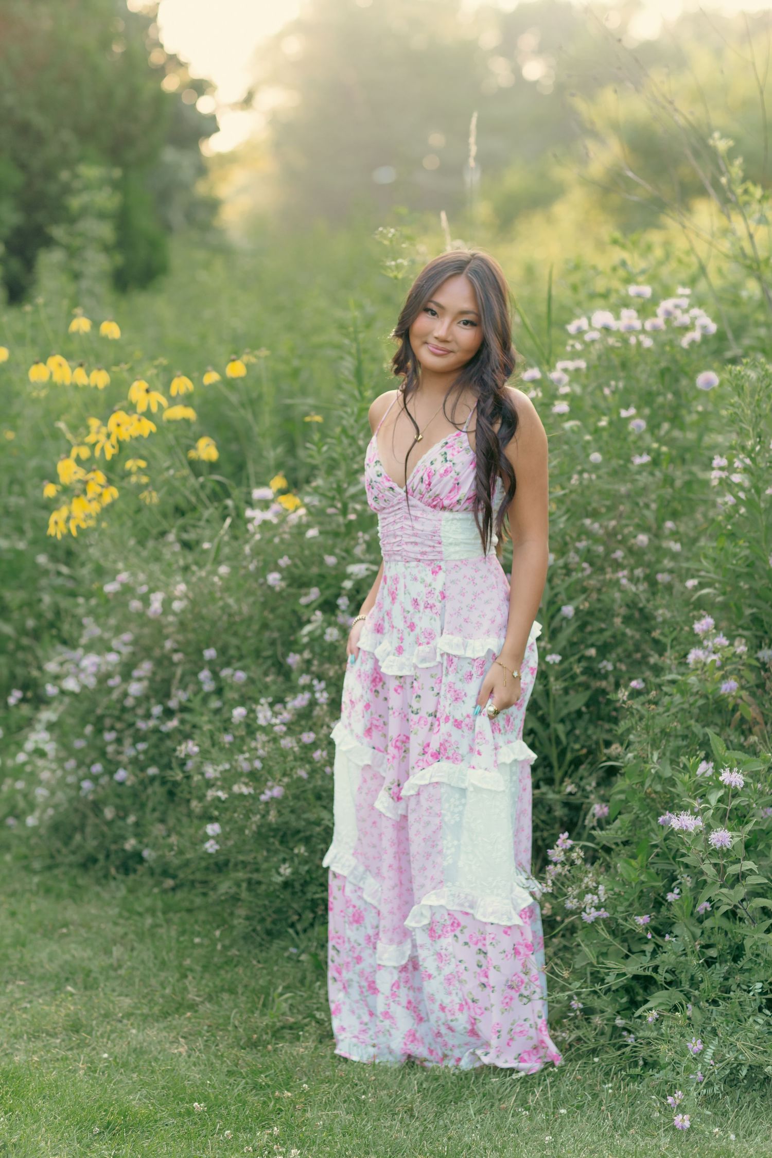 Young woman in pink floral maxi dress posing among wildflowers in a lush green garden at golden hour.