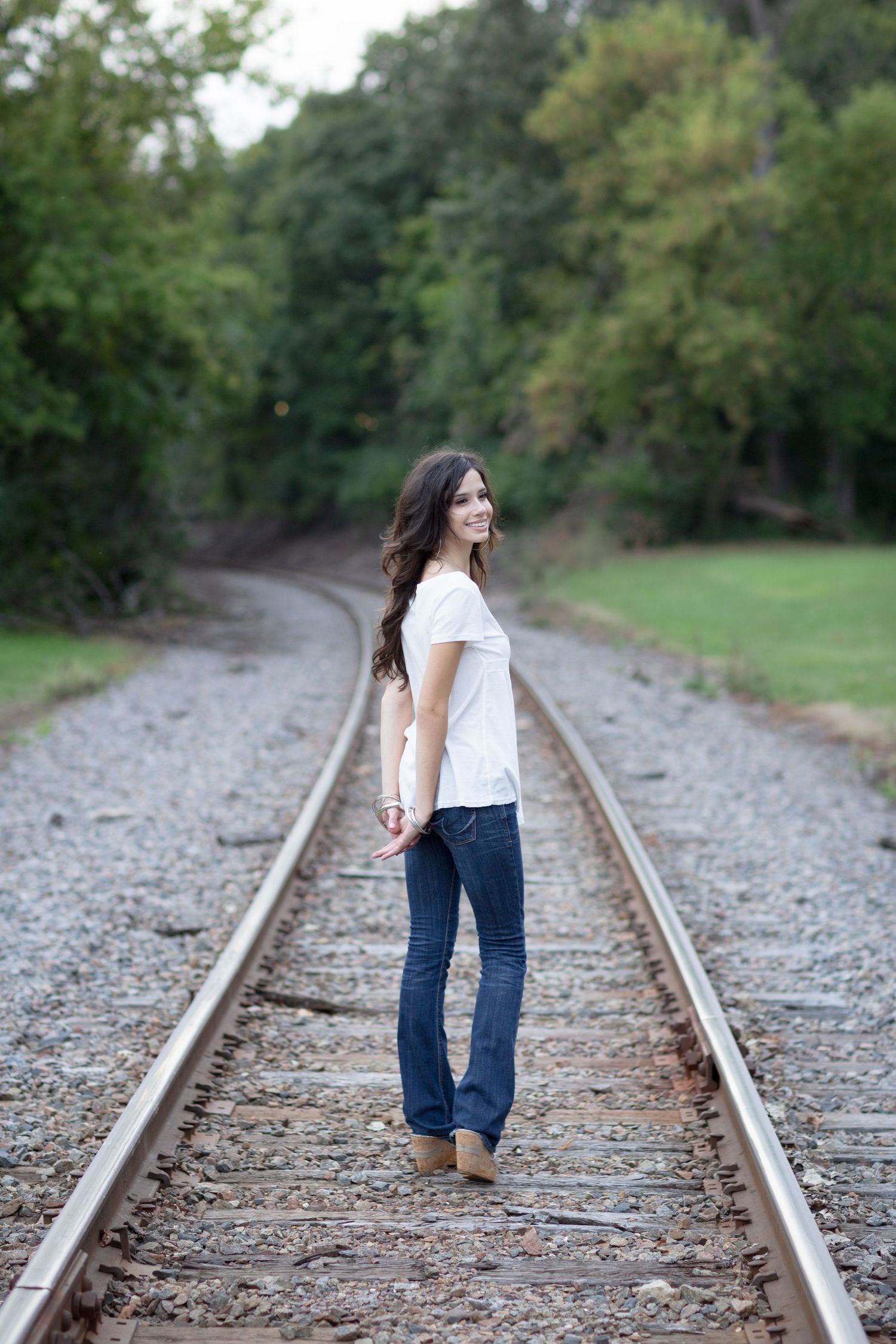 Young woman in white top and jeans smiling while standing on railroad tracks surrounded by green trees.