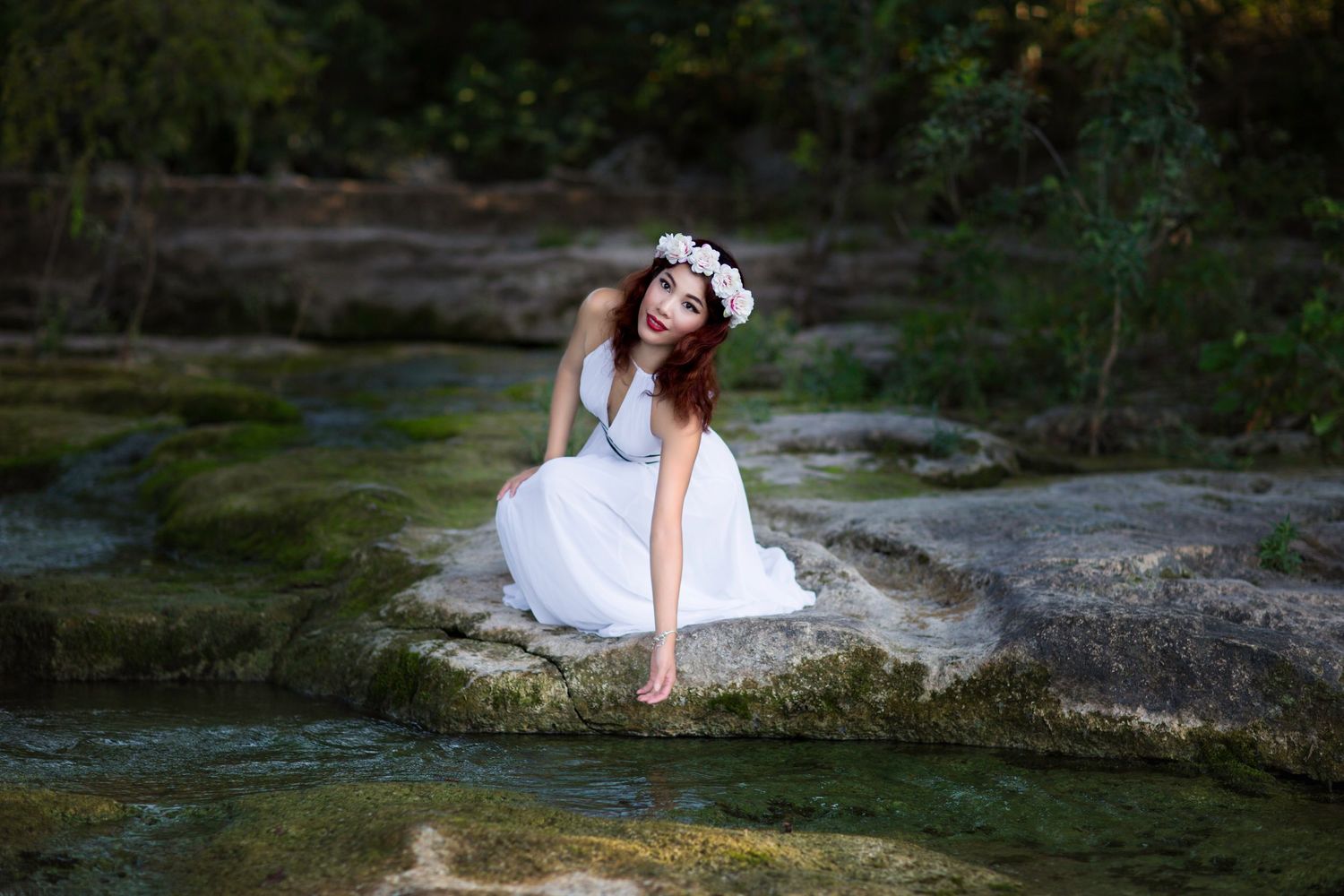 A young woman in a white dress and floral crown sits gracefully on mossy rocks beside a serene woodland stream.
