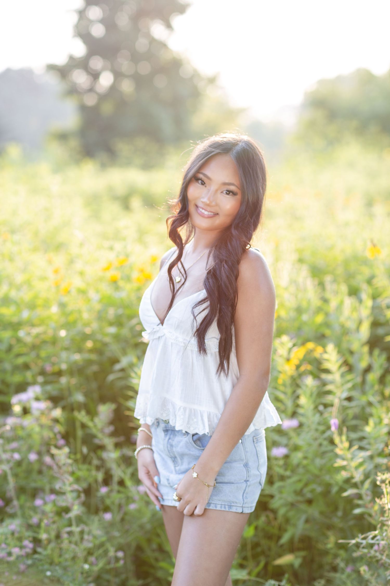 Smiling young woman in white top and denim shorts posing in a sunlit wildflower field.