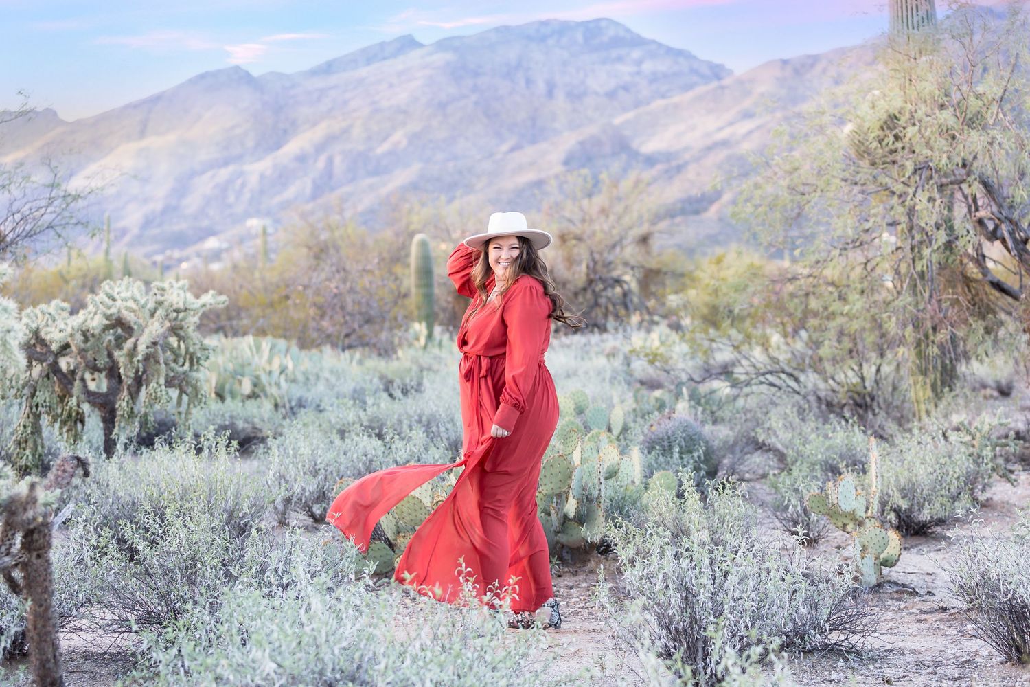 Woman in flowing red dress and white hat posing in desert landscape with mountains behind.