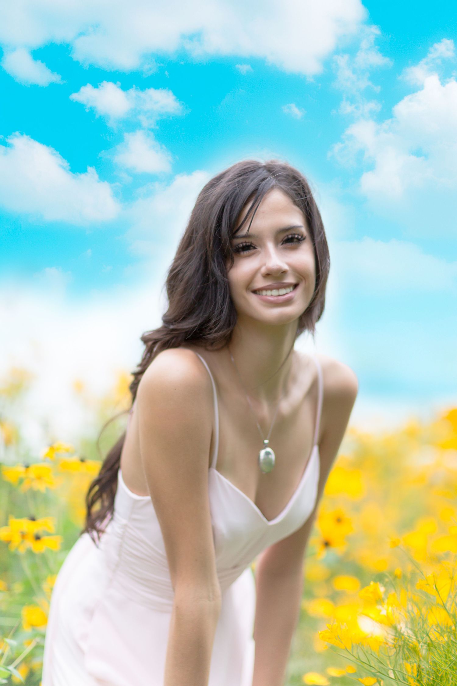 Smiling young woman in white dress among yellow wildflowers under blue sky with clouds.