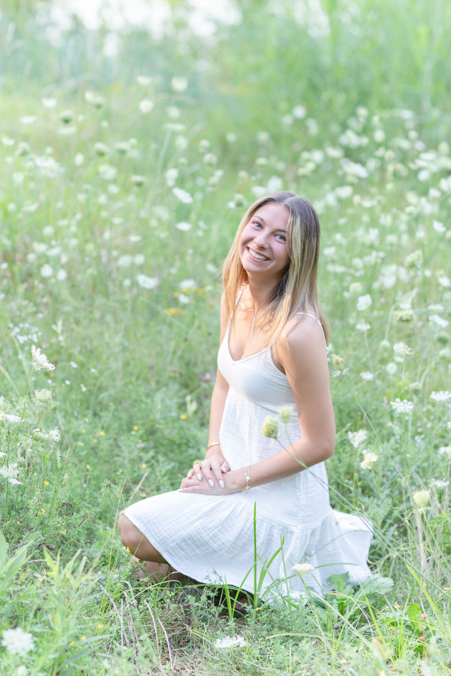 Young woman in white sundress sitting in a lush green meadow with white wildflowers, smiling joyfully.
