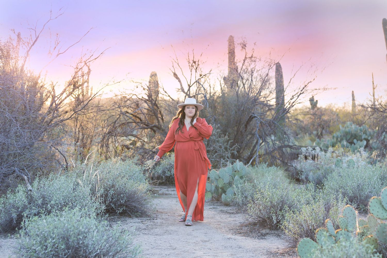Pregnant woman in red dress and white hat walking among desert cacti at sunset.