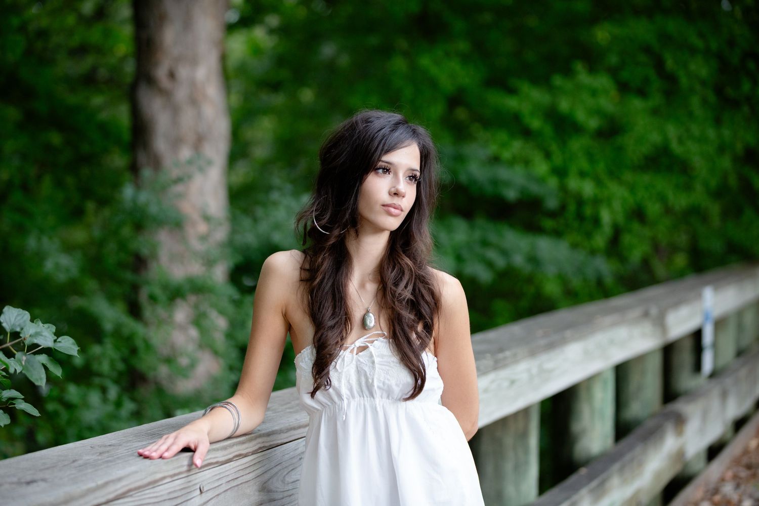 Young woman in white dress leaning on wooden bridge railing surrounded by lush green trees.