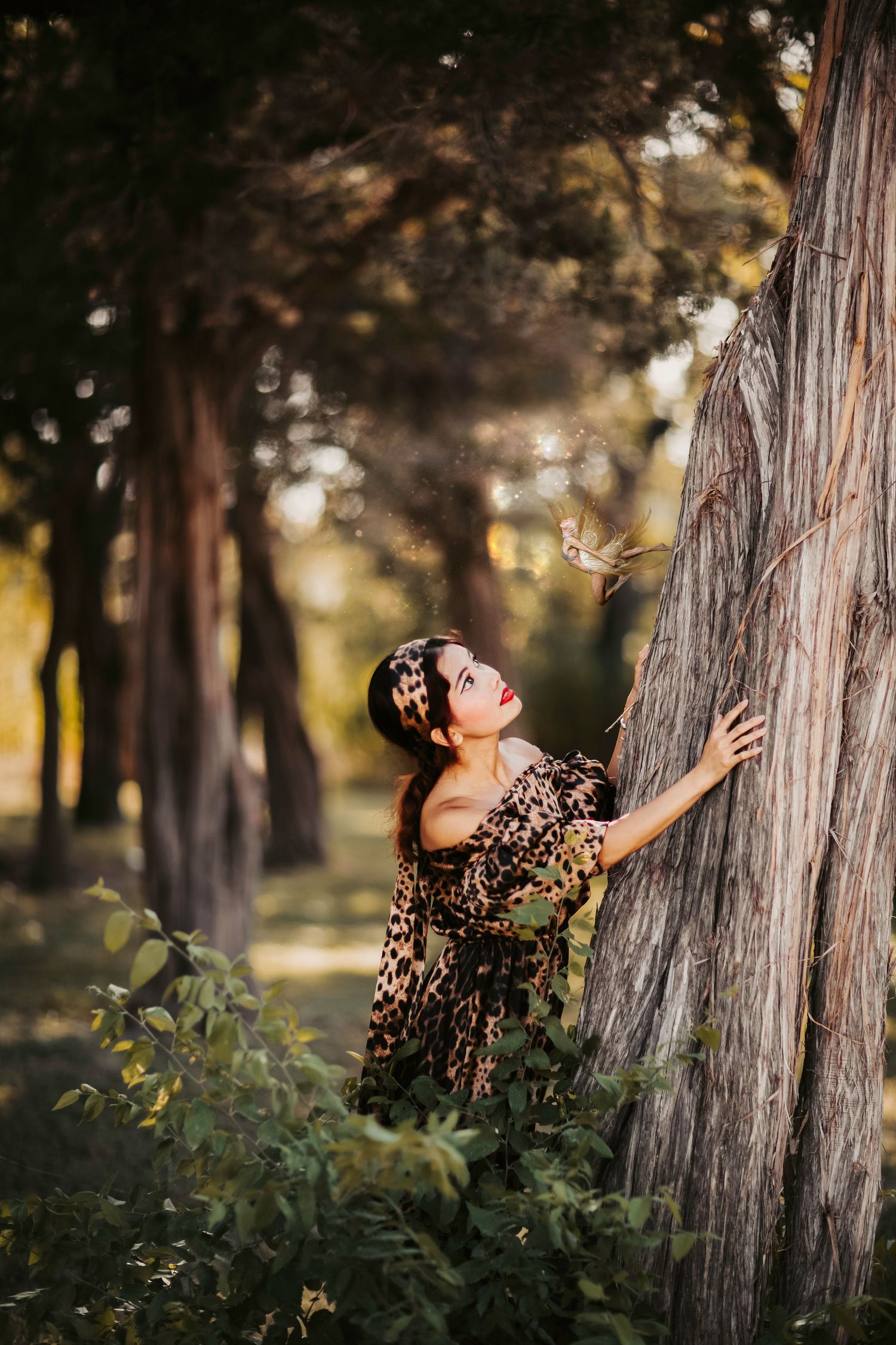 Woman in leopard print dress touching tree bark in sunlit forest with bokeh background. Fine Art Portrait Photography whimsical