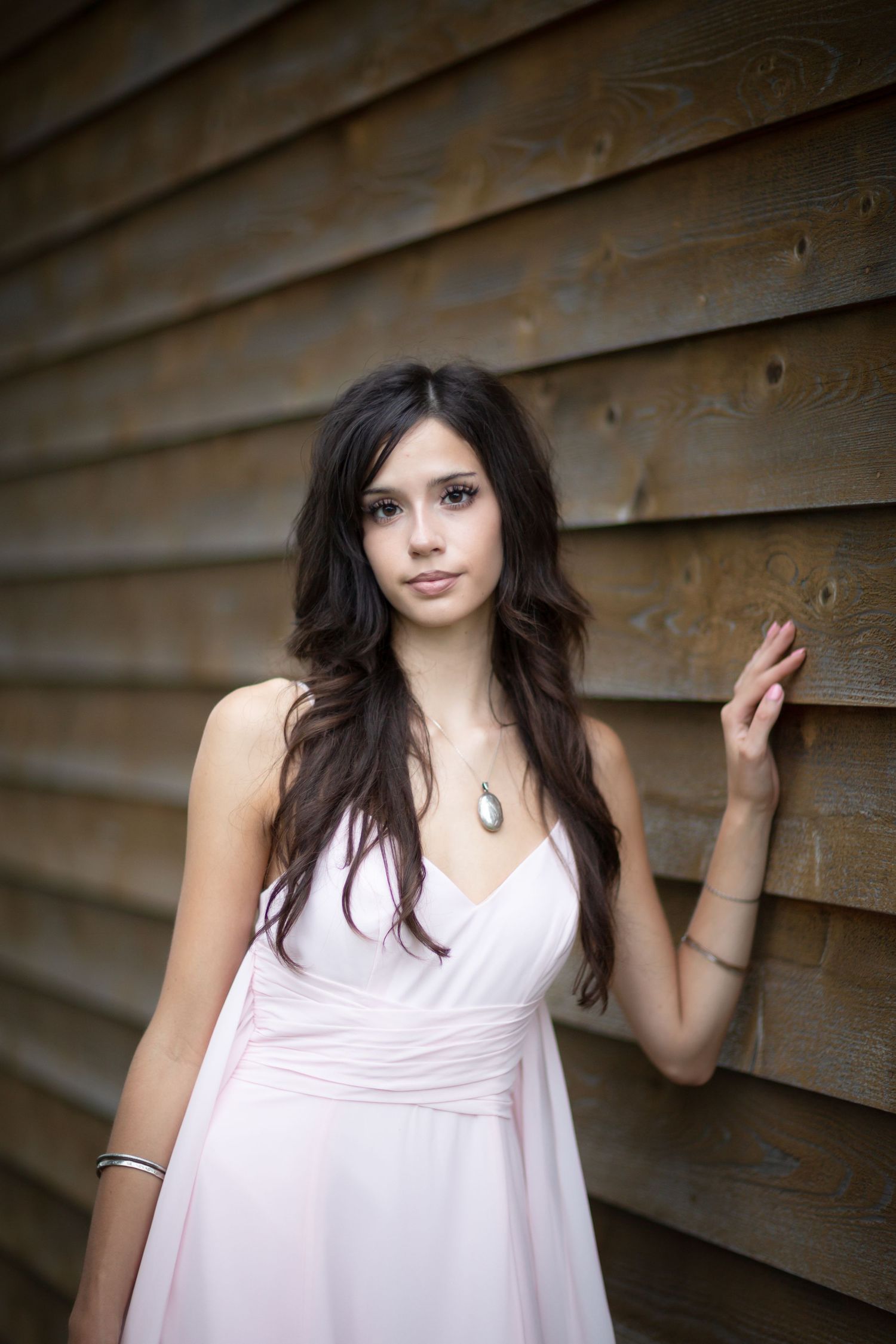 Young brunette woman in pink dress leaning against rustic wooden wall wearing pendant necklace.