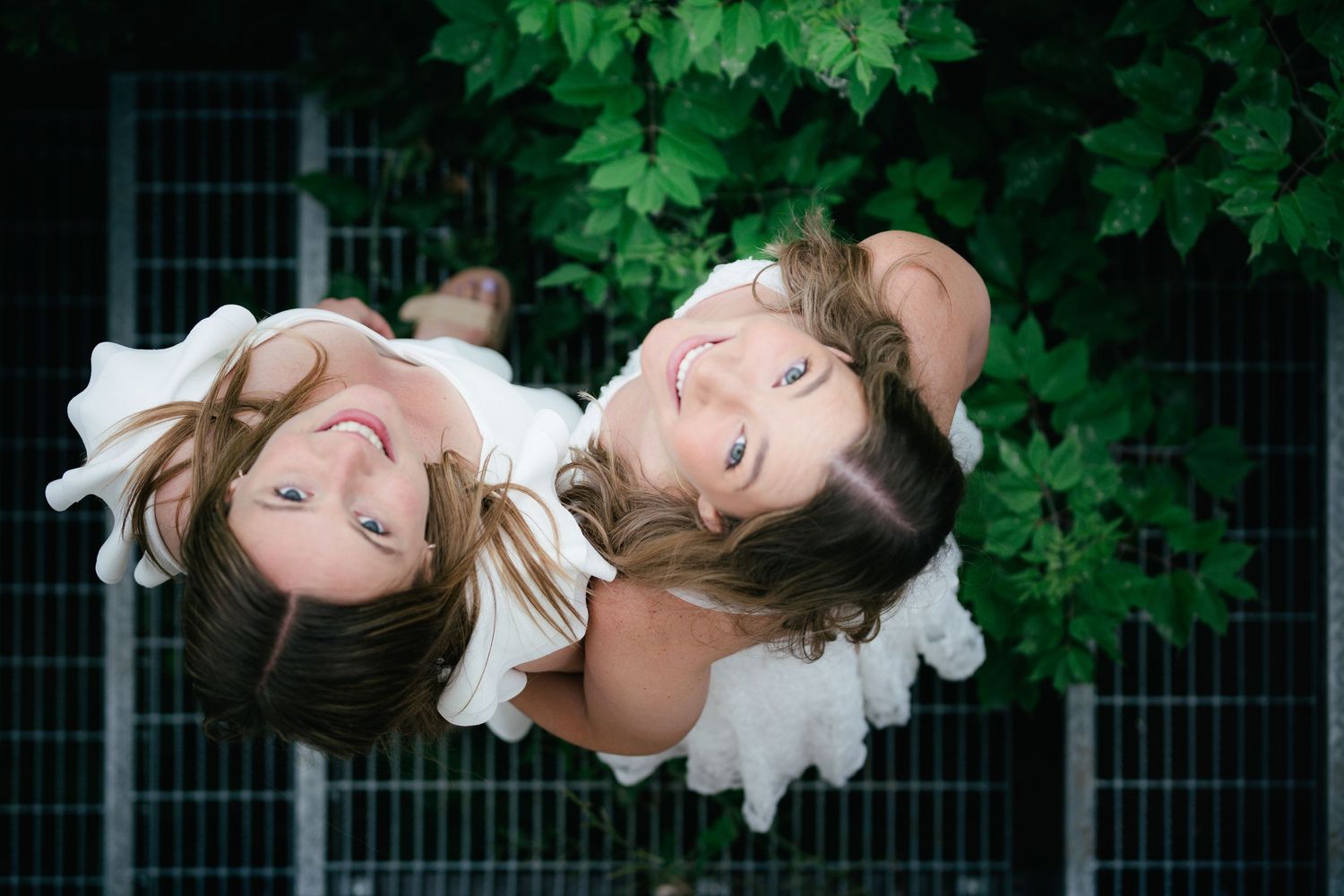 Two smiling girls in white dresses pose upside down surrounded by lush green leaves outdoors.