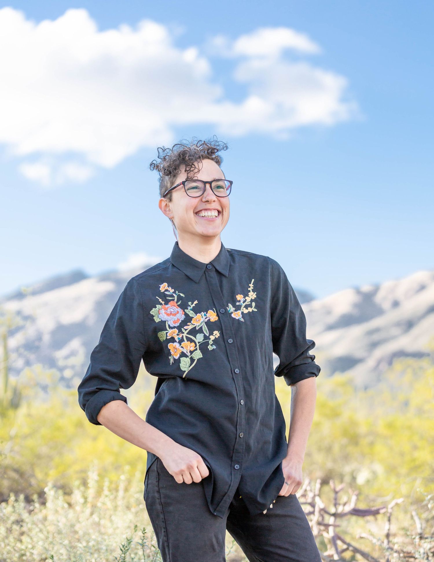 Smiling person with curly hair and glasses wearing a floral embroidered black shirt outdoors.