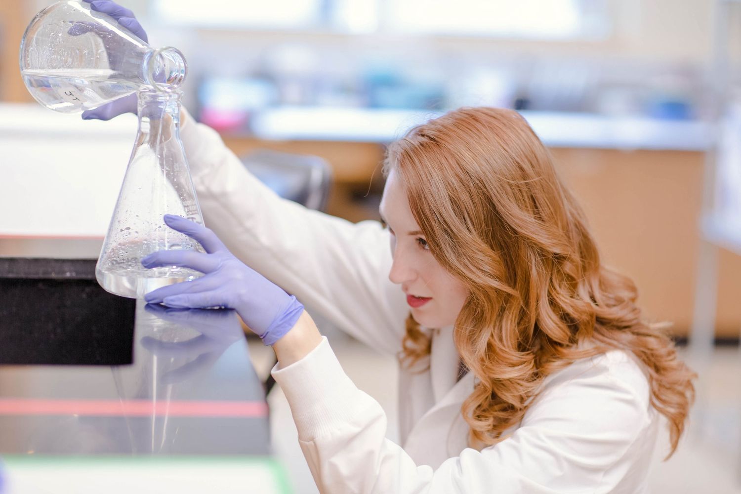 Female scientist in white lab coat and blue gloves examining flask in laboratory setting.