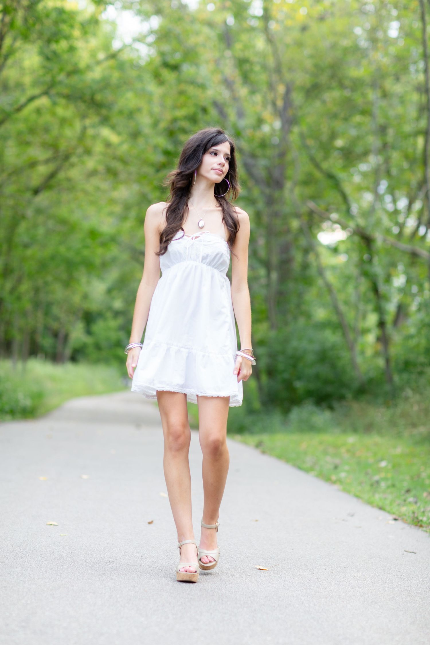 Young woman in white sundress walking on a tree-lined path in a lush green park on a summer day.
