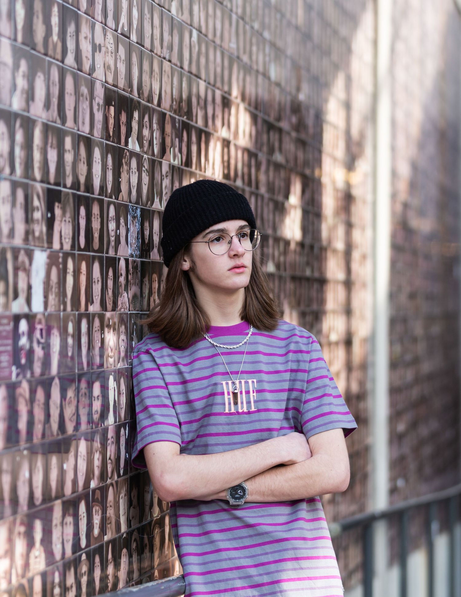 Teen boy with glasses and beanie leaning against brick wall covered in photos, arms crossed.