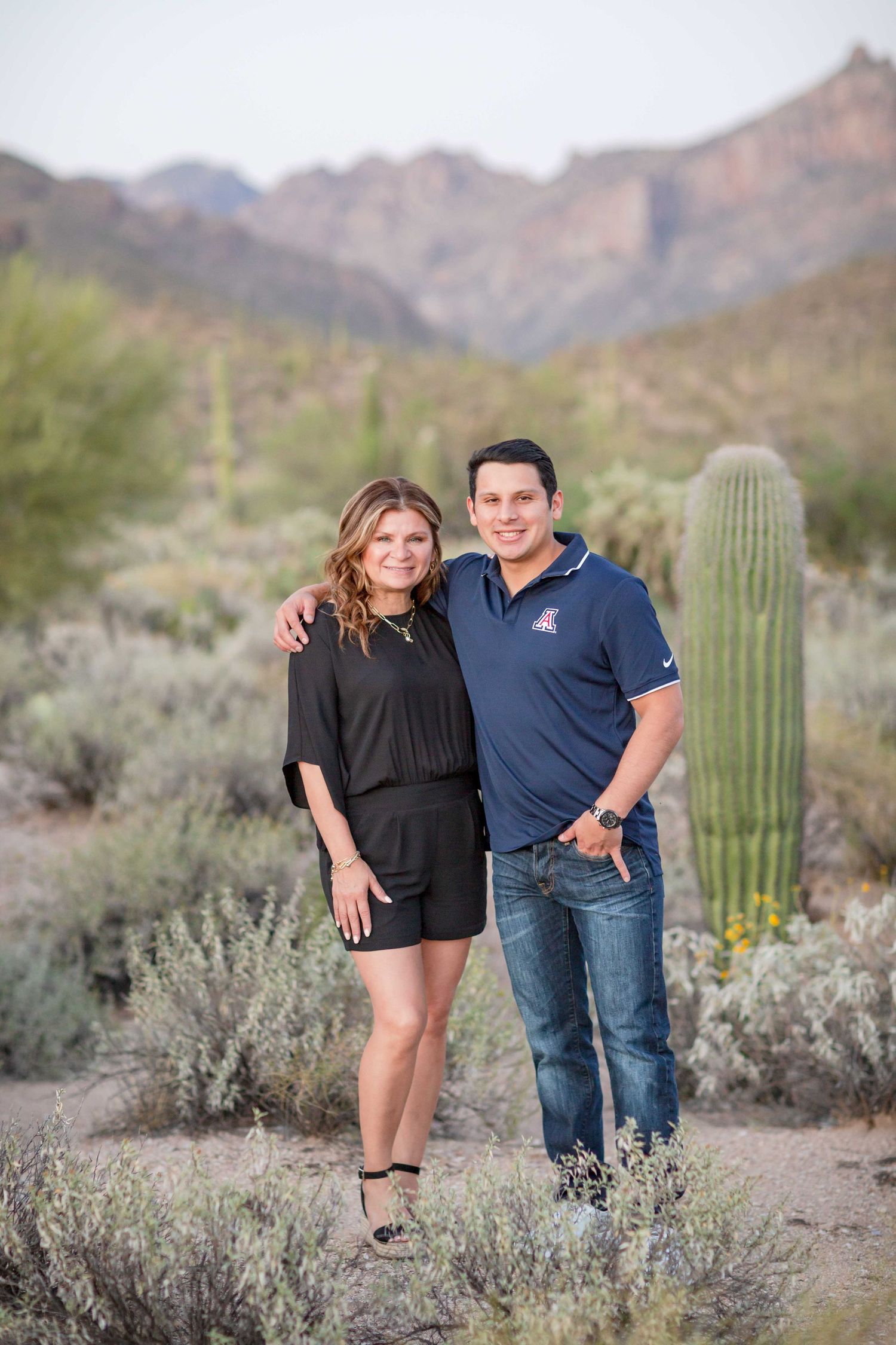 Smiling couple posing together in Arizona desert landscape with saguaro cacti and mountains.