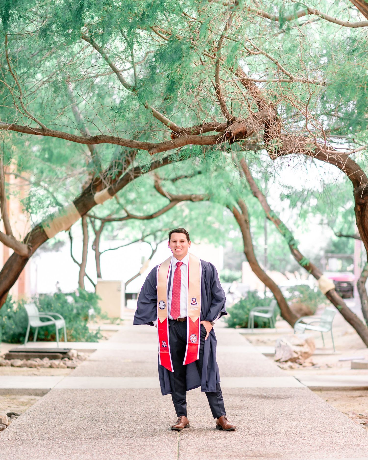 Graduate in cap and gown with red stole posing under trees on a campus walkway.