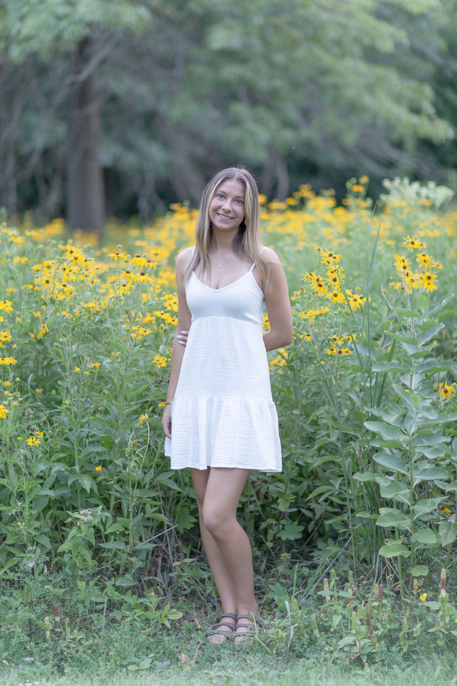 Young blonde woman in white sundress smiling among yellow wildflowers in a lush green garden.
