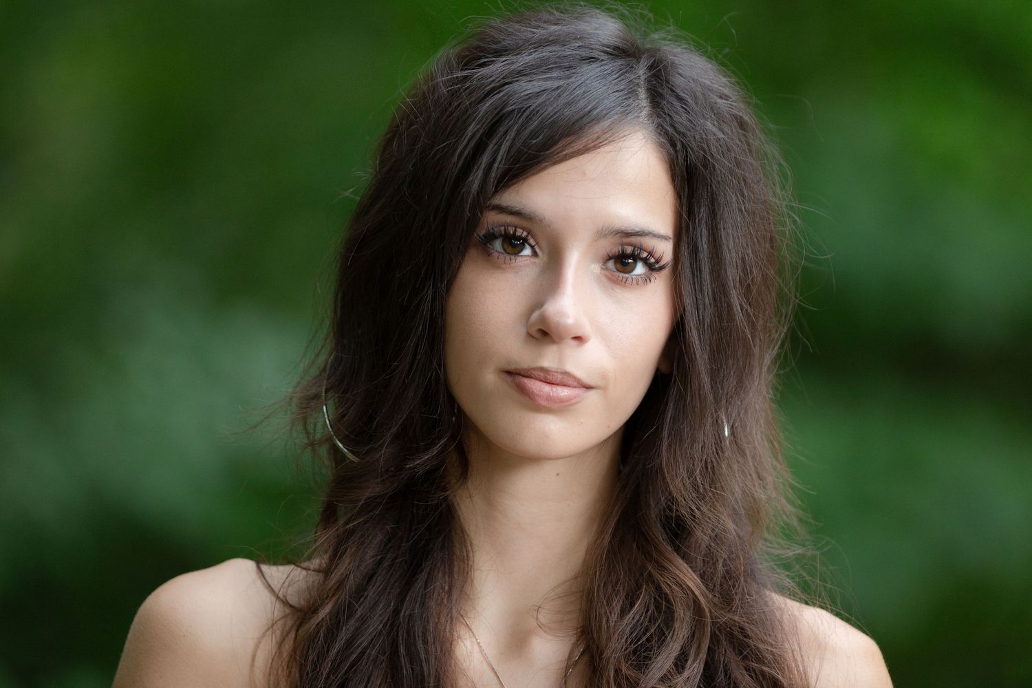 Young woman with long dark hair posing outdoors with a natural green bokeh background.