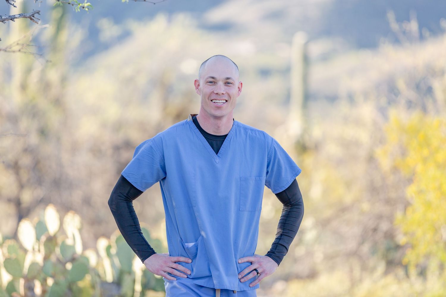 Man in blue medical scrubs standing confidently outdoors in desert setting with cacti background.