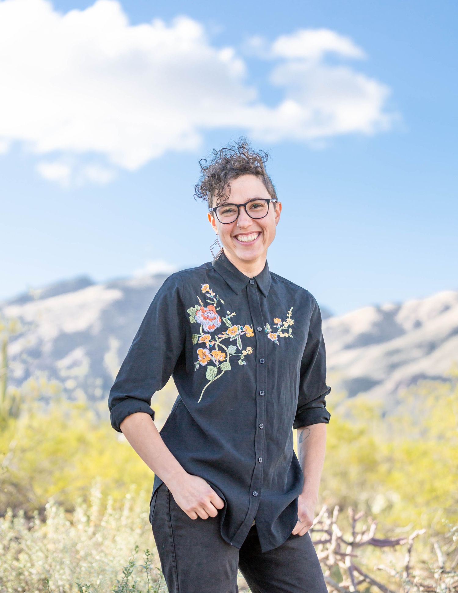 Smiling woman in floral embroidered denim shirt posing outdoors with mountain backdrop.