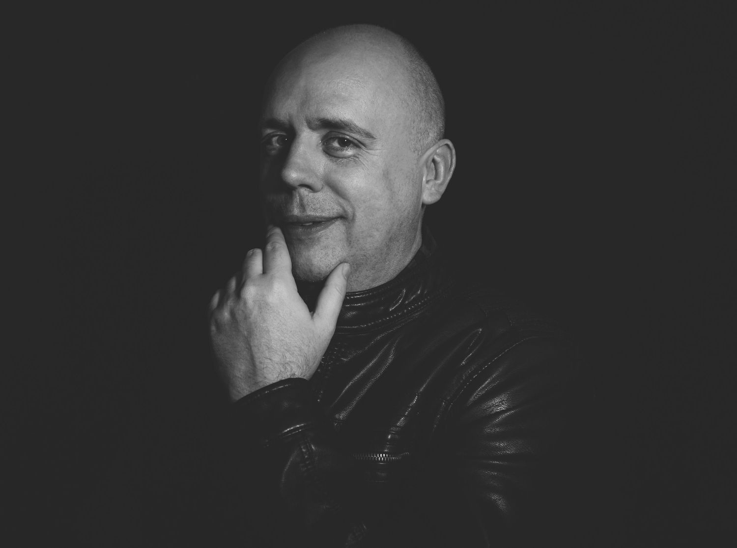 Bald man in black leather jacket posing thoughtfully with hand on chin against dark background.