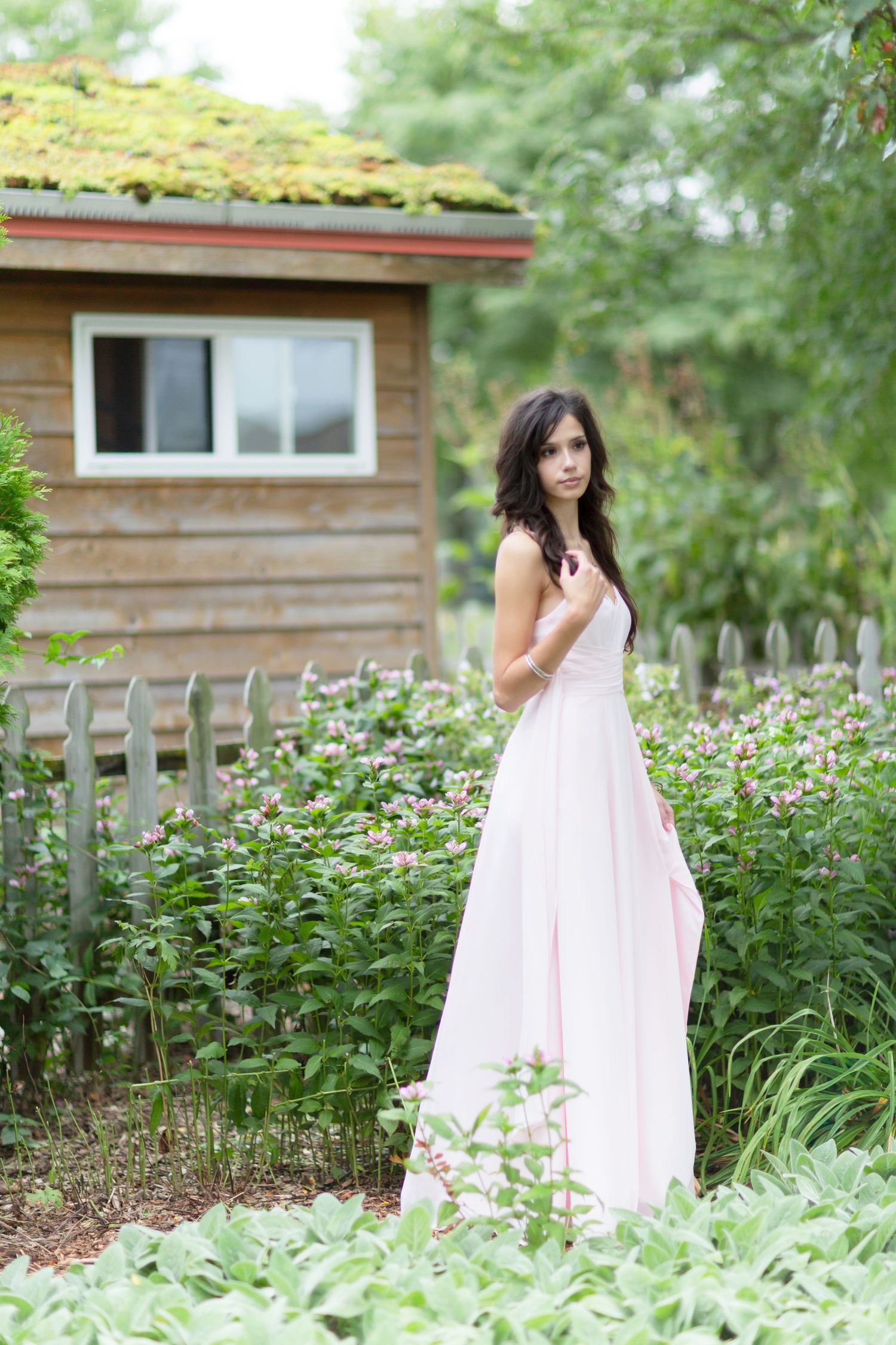 Young woman in pink gown standing among garden flowers near wooden cottage with green roof.