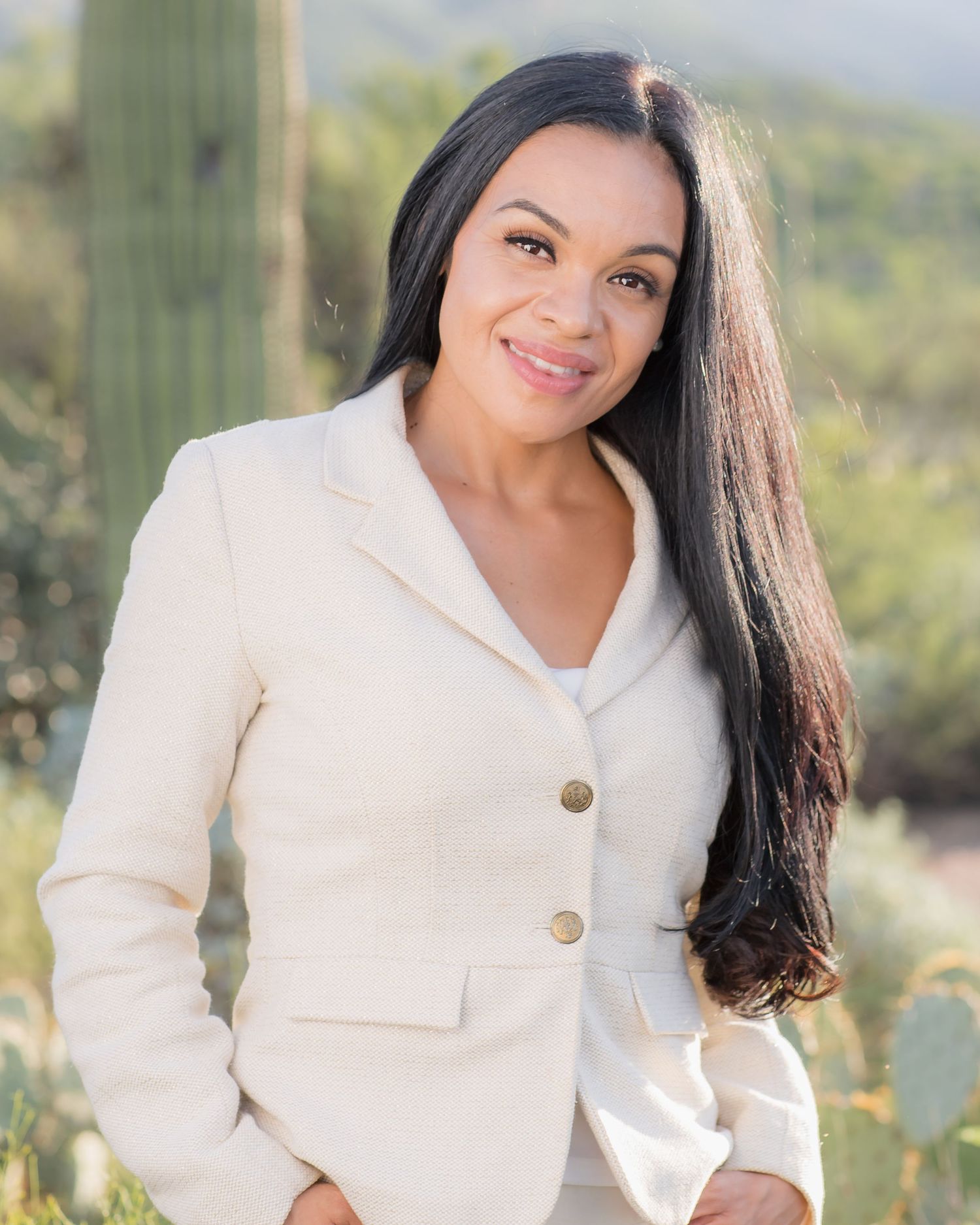 Professional woman in cream blazer with long black hair posing near saguaro cacti outdoors.