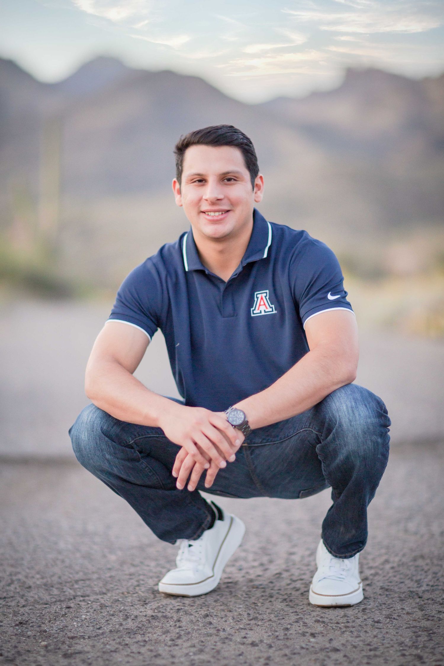 Young man in University of Arizona polo shirt crouching on a road with mountains behind.