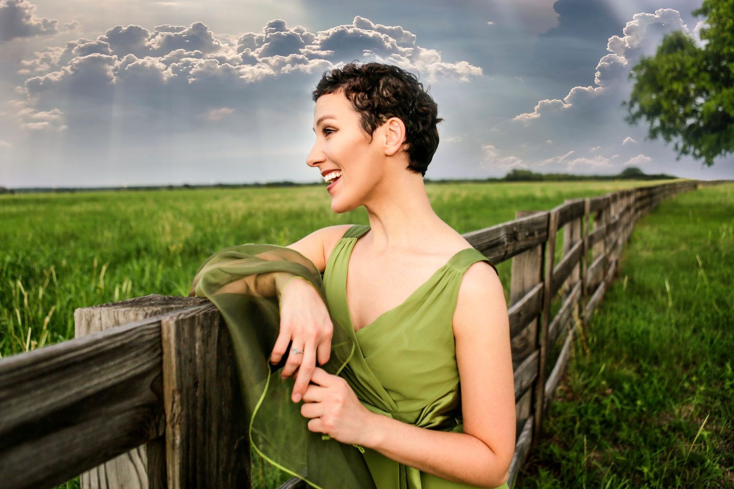 Woman in green dress leaning on wooden fence in lush field, smiling under dramatic cloudy sky.