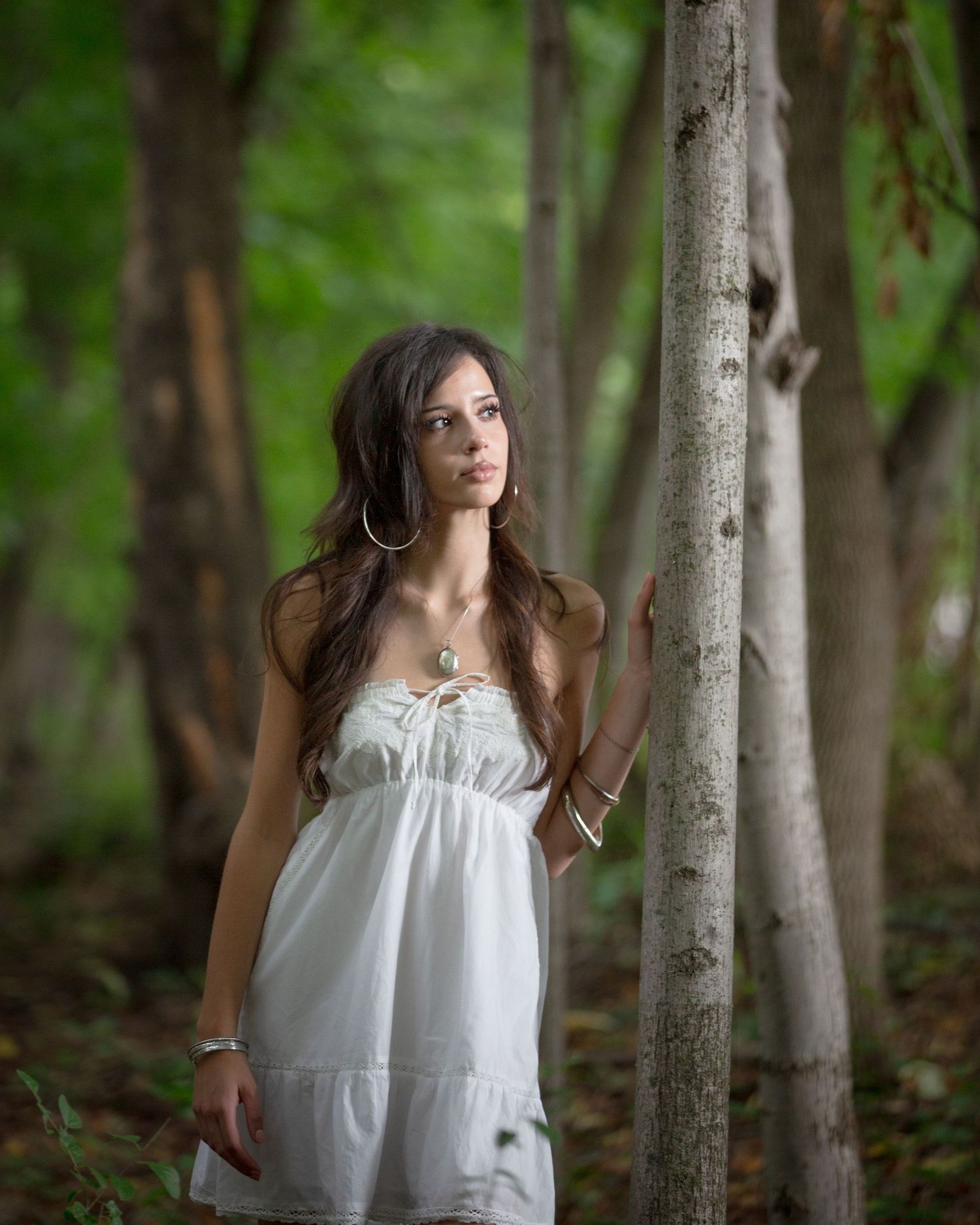 Young woman in white sundress standing among trees in a lush green forest, wearing hoop earrings and bracelets.