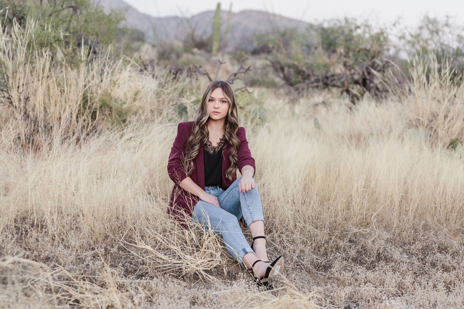 Young woman in a burgundy blazer and ripped jeans sits in a dry grassy desert landscape.