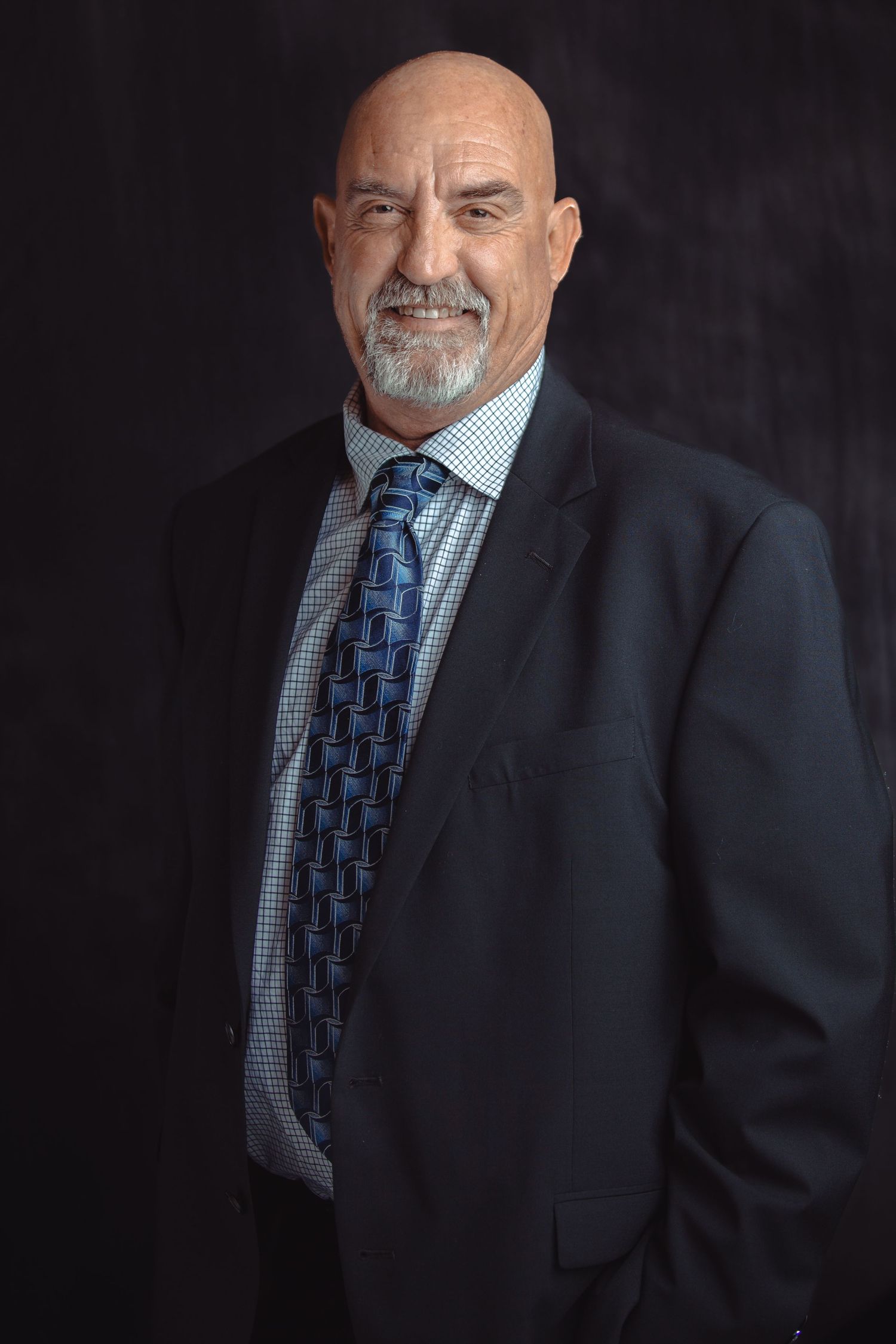 Bald man with gray beard in dark suit and blue tie posing against a dark background.