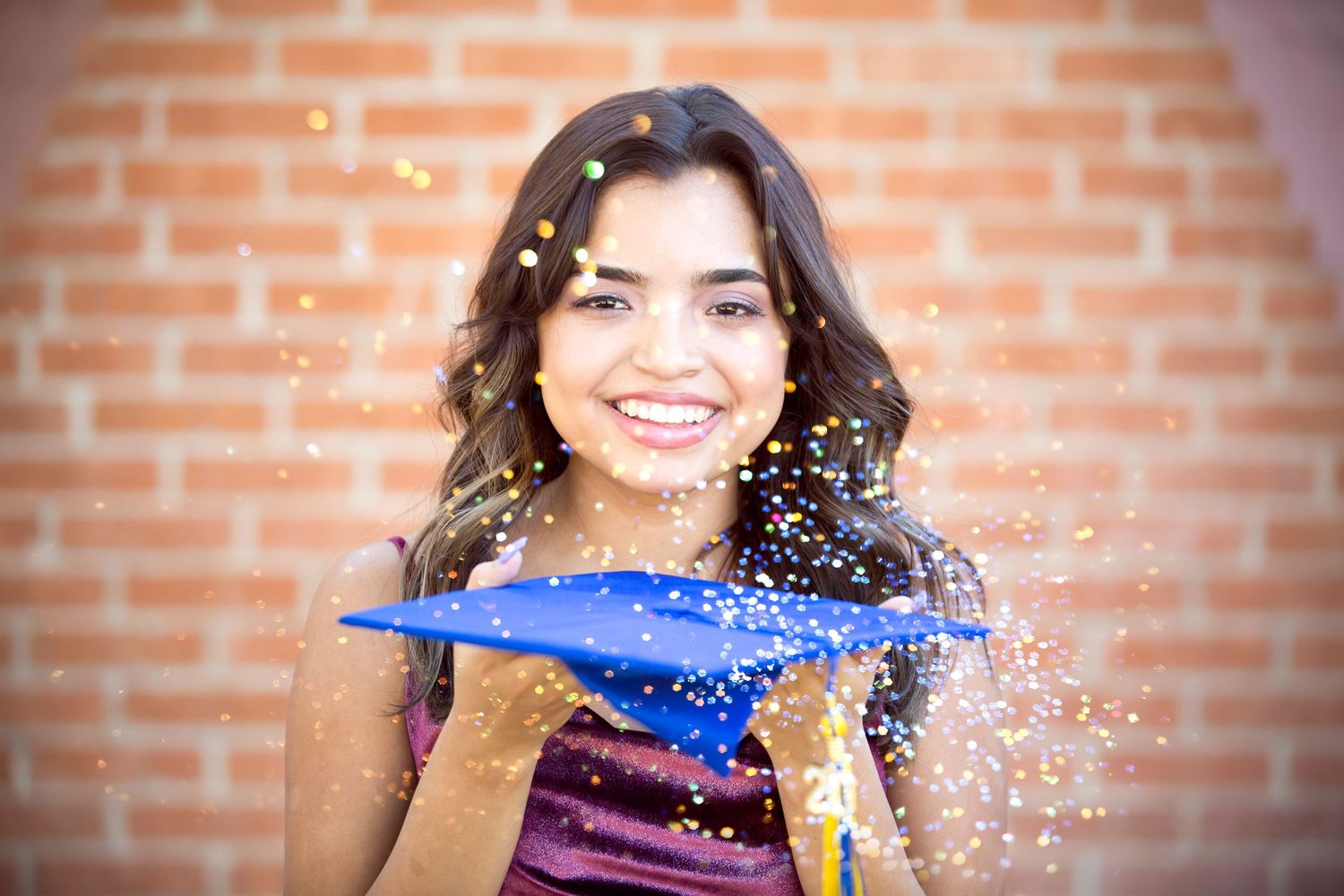 Smiling young woman in graduation cap blowing colorful confetti against brick wall background.