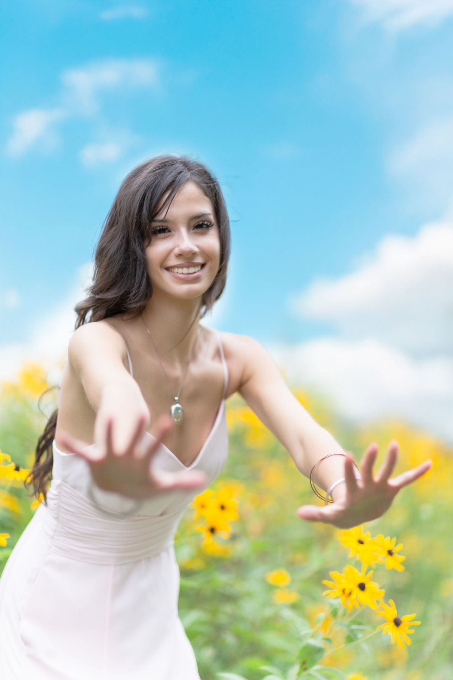 Happy young woman in white dress reaching toward camera in a field of yellow sunflowers under blue sky.