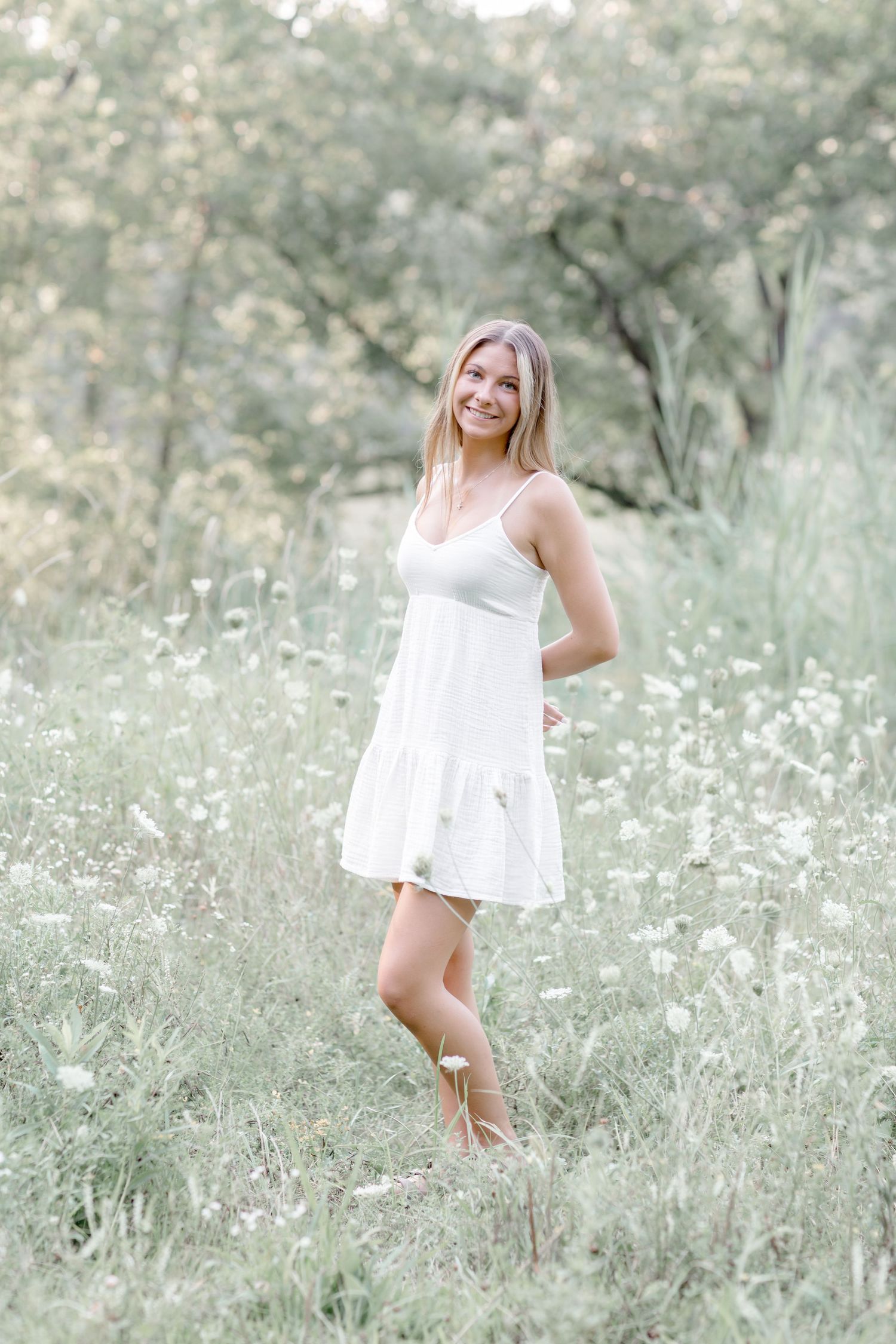 Smiling young woman in white sundress standing in wildflower meadow with lush green trees in background.