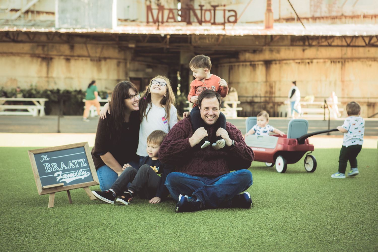 Family of five posing playfully on green turf near a Magnolia sign with a chalkboard.