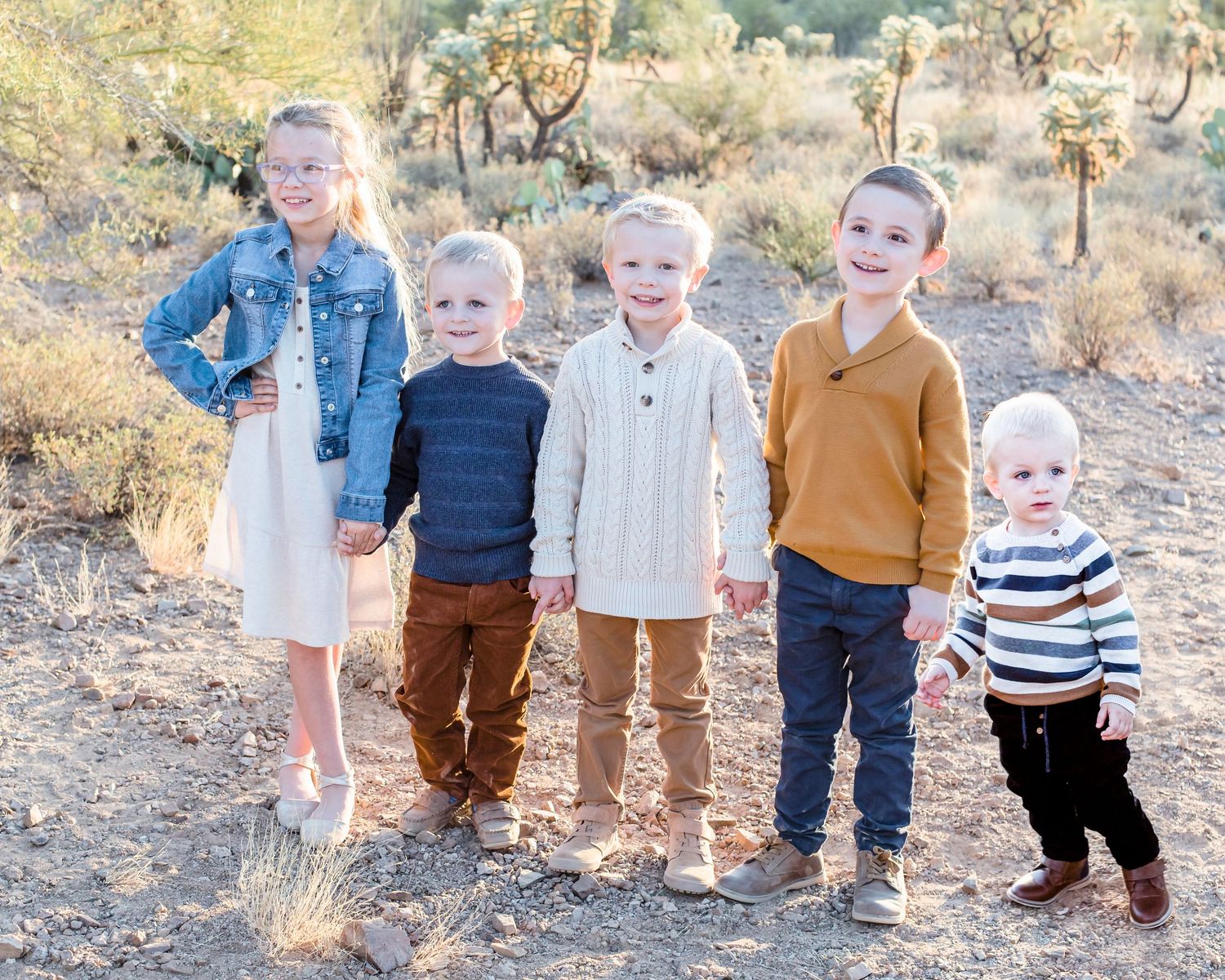 Five young children holding hands smiling outdoors in desert landscape with cacti background.