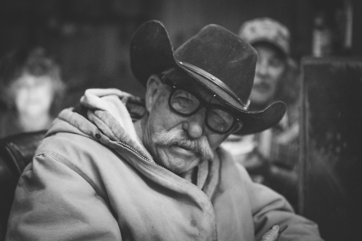 Close-up black and white portrait of older man in dark cowboy hat and glasses looking down.