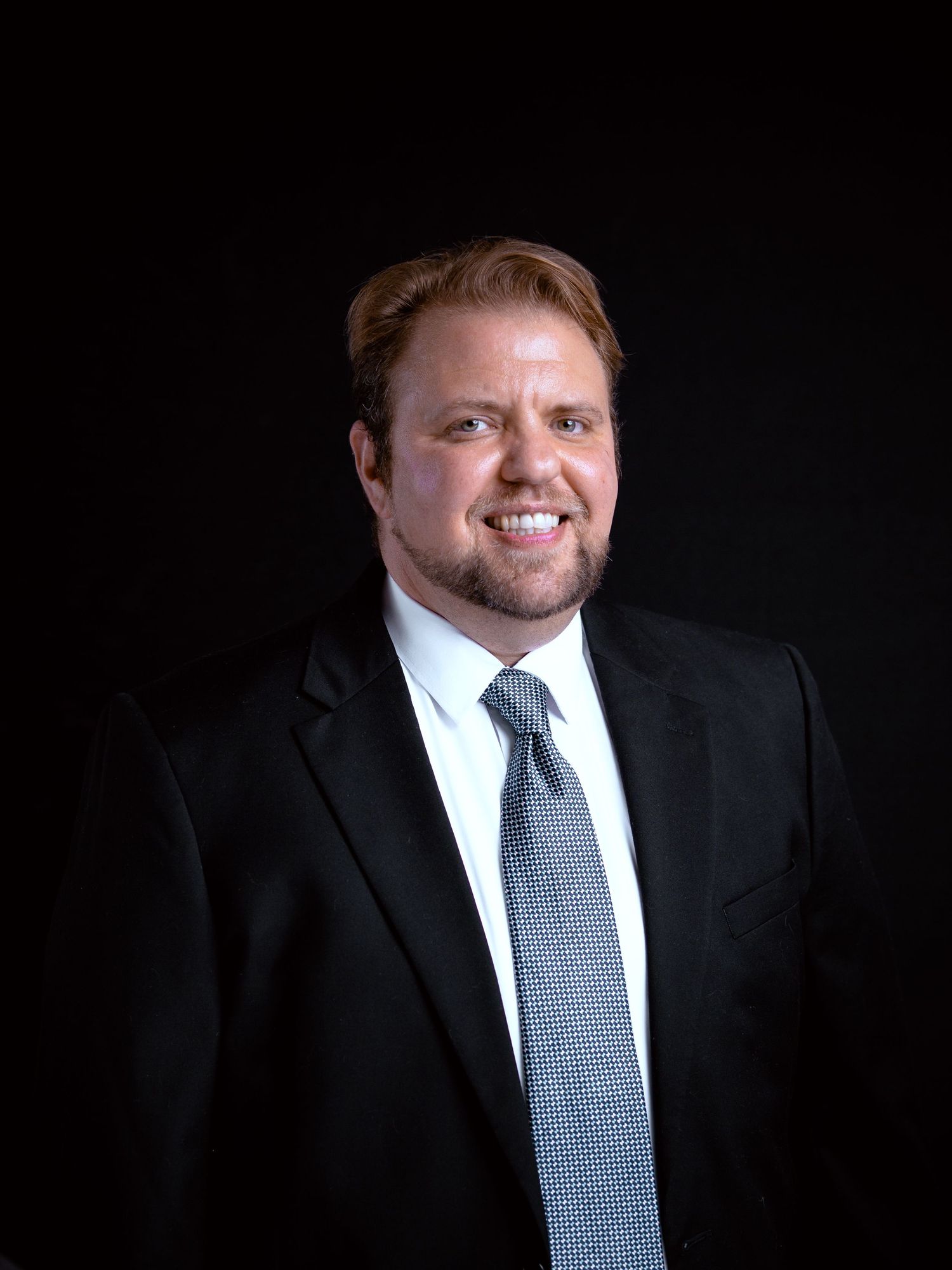 Professional headshot of smiling man in black suit and gray tie against dark background.