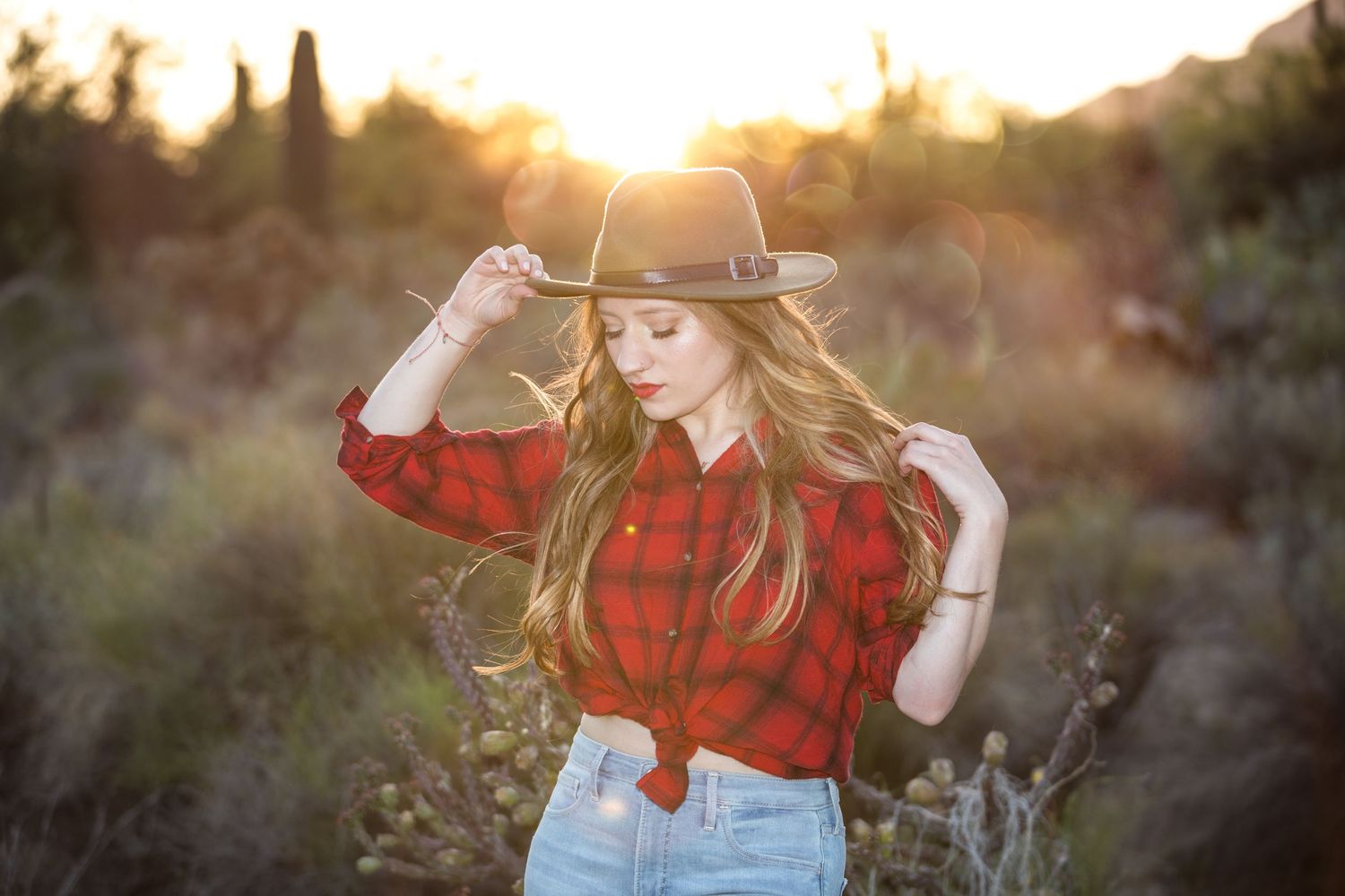Young woman in red plaid shirt and wide-brim hat posing at golden hour in desert field.