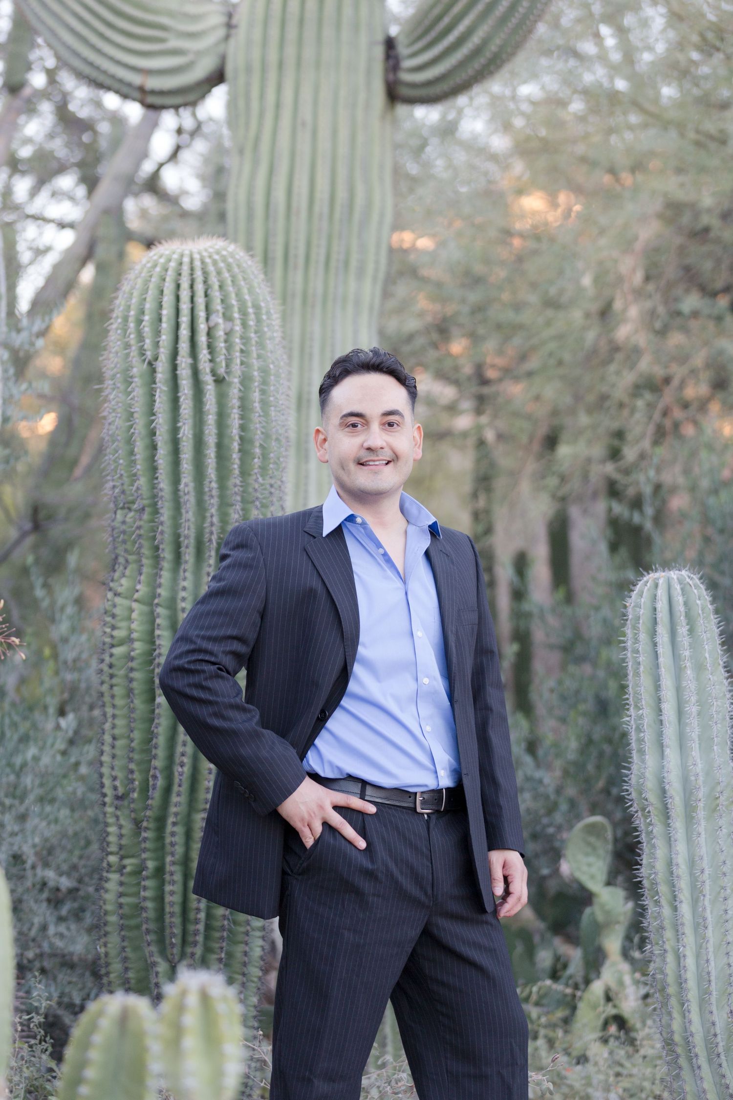 Smiling man in dark pinstripe suit posing among tall saguaro cacti in desert setting.