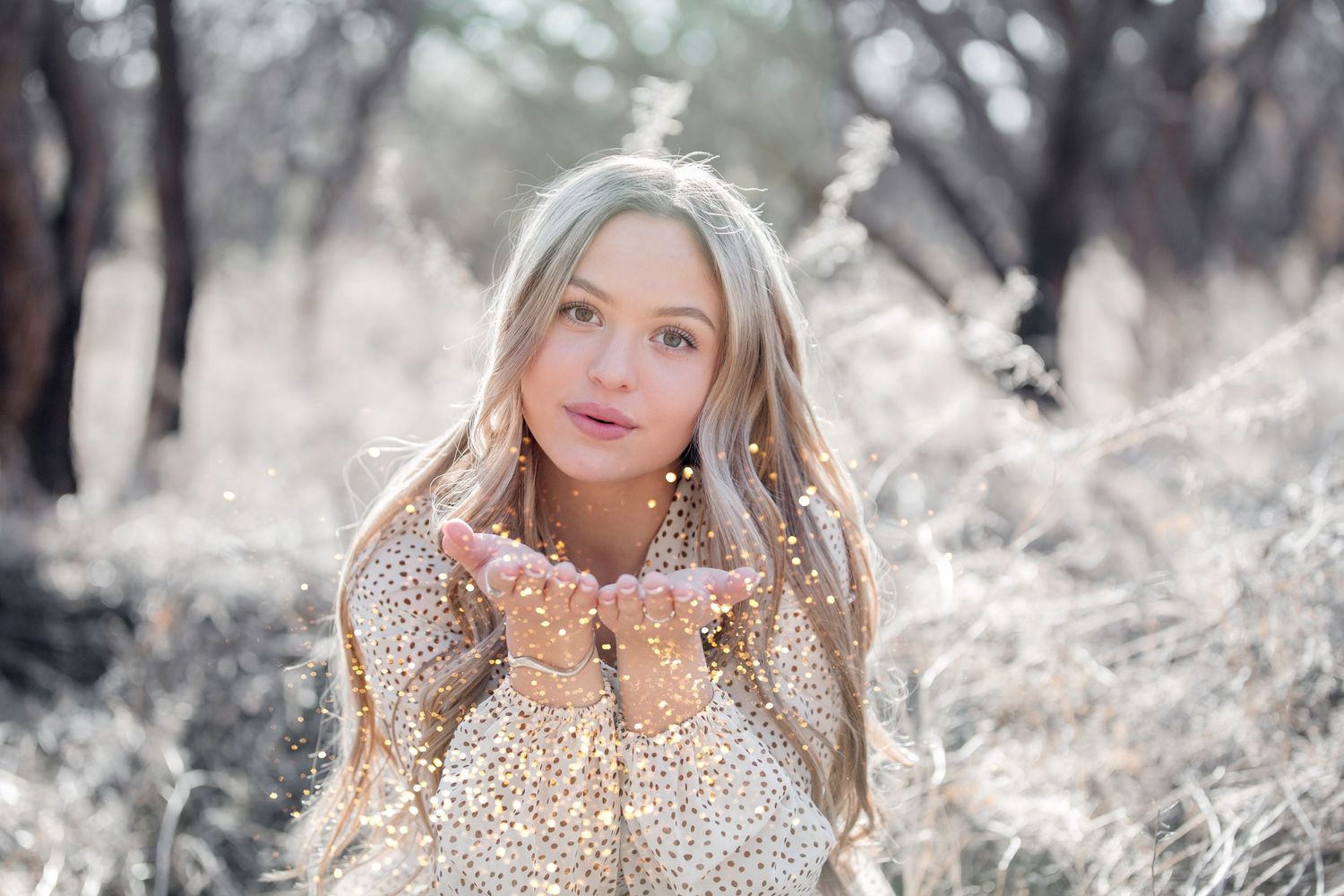Young blonde woman blowing glitter from her hands outdoors in a dreamy winter forest setting.