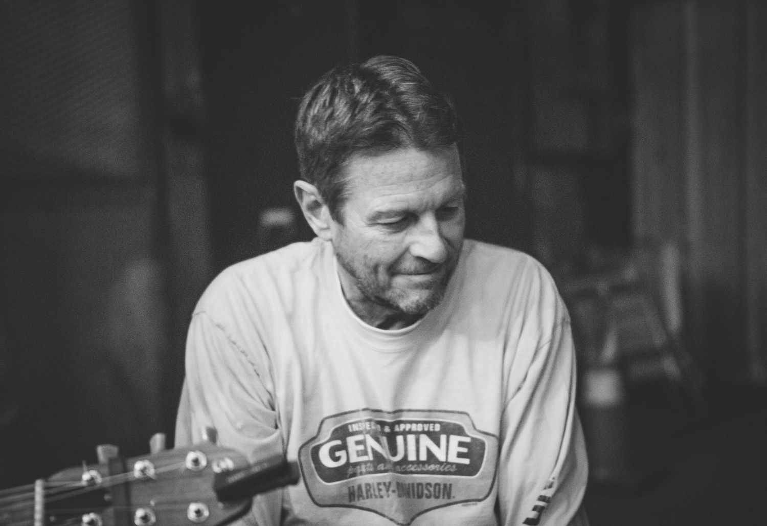 Man in Harley Davidson shirt smiling while playing acoustic guitar in black and white photo.