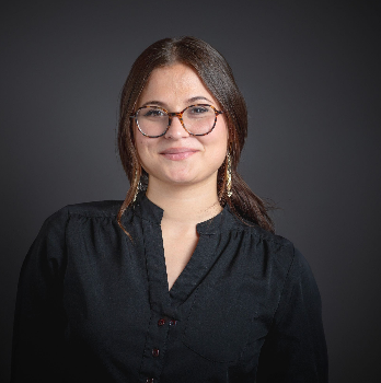A young woman with tortoiseshell glasses and gold earrings poses in a black blouse against a dark gray backdrop.