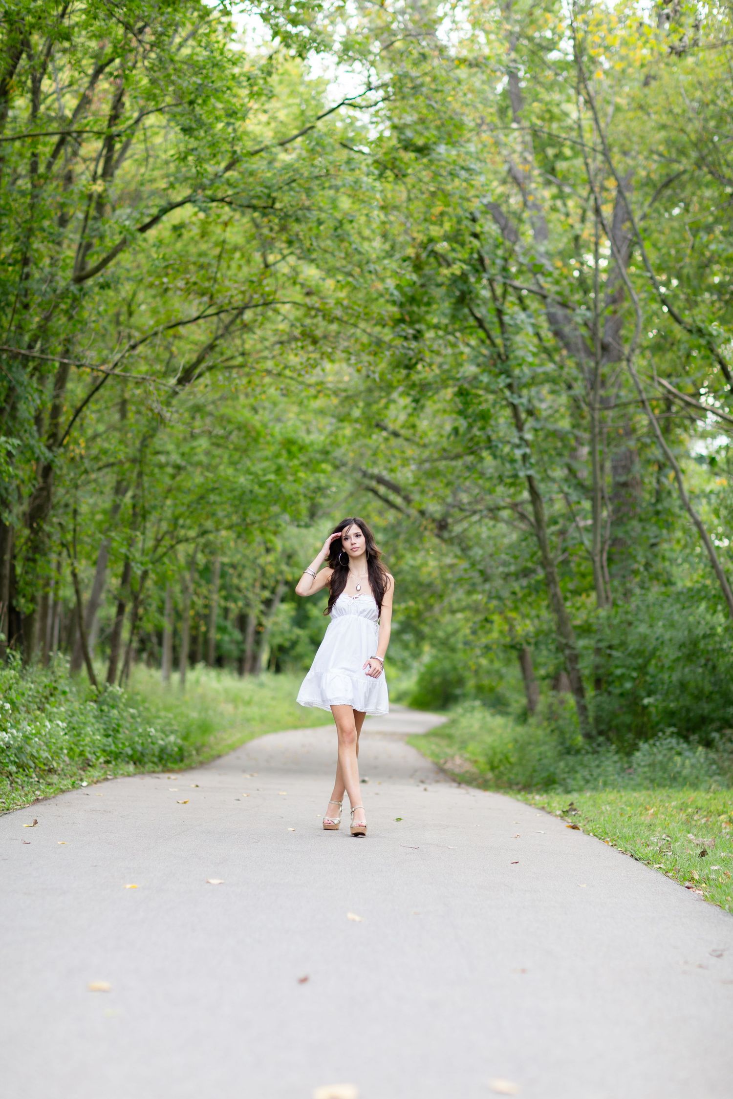 Young woman in white dress walking along tree-lined path in lush green park on a summer day.