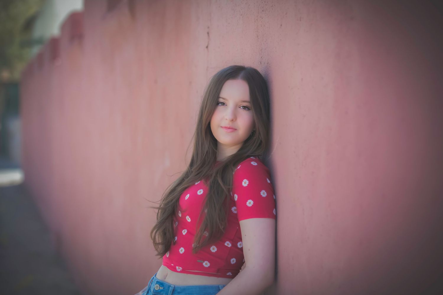 Young brunette girl in red polka dot top leaning against pink wall outdoors.