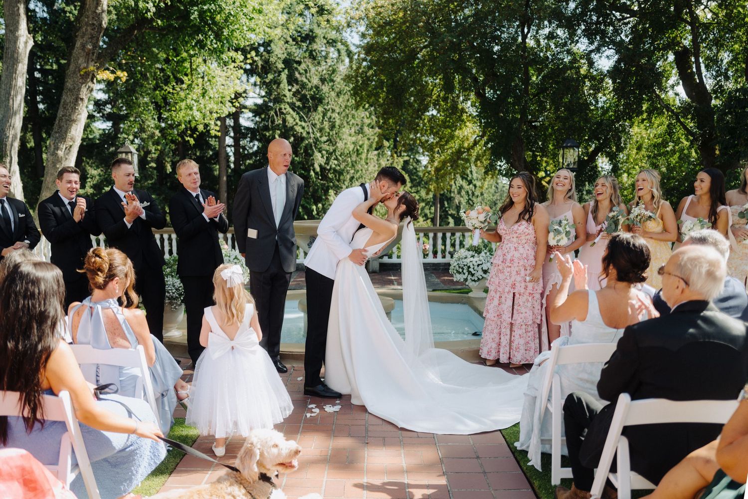 An outdoor wedding ceremony takes place under a canopy of trees with guests seated on white chairs.