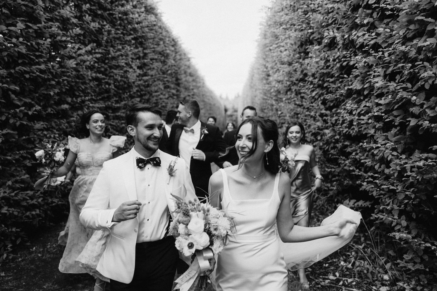 Black and white photo of wedding celebration with guests walking through a hedge-lined path.