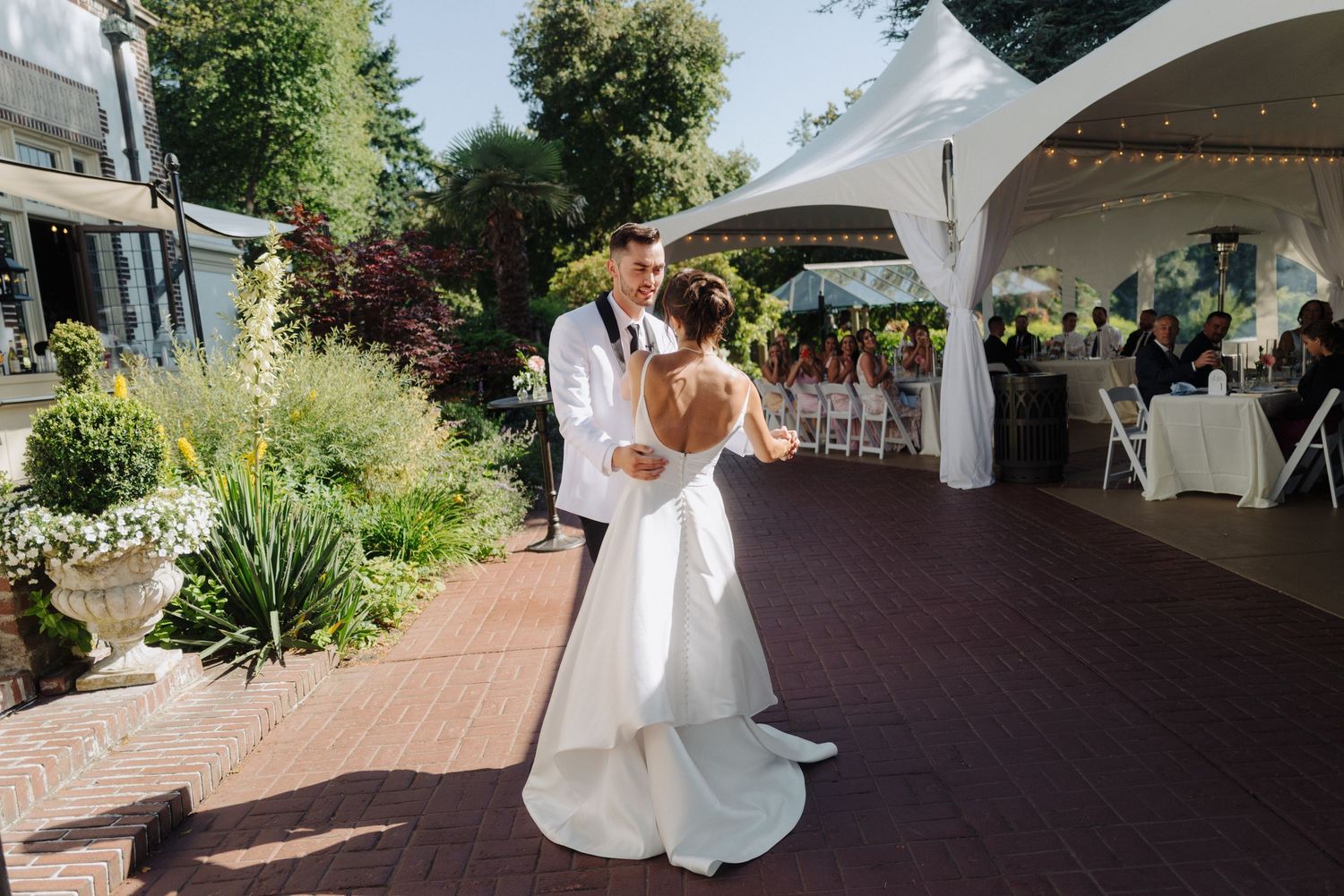 White wedding arch and chairs arranged on a paved outdoor ceremony space surrounded by greenery.