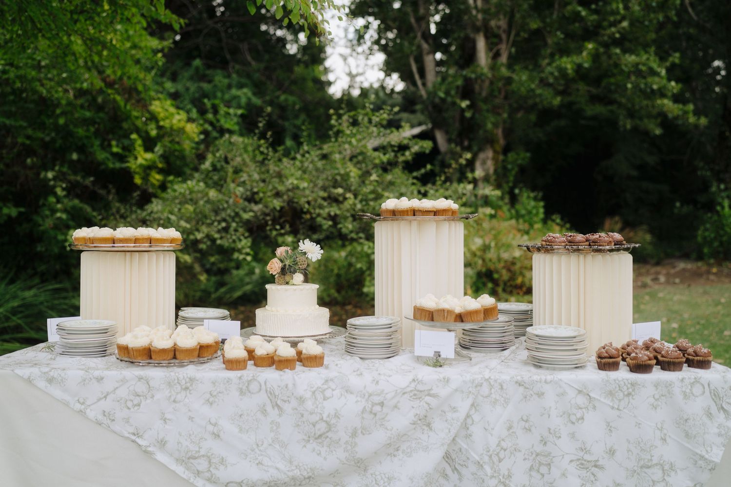 Elegant dessert table with white tablecloth displays wedding cakes and cupcakes in an outdoor garden setting.