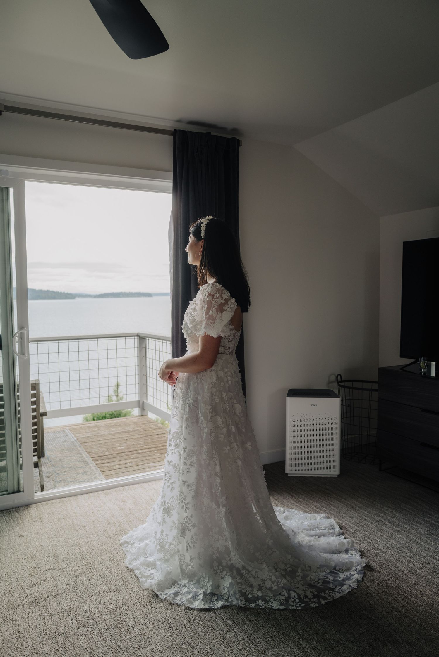 A beautiful lace wedding dress displayed in a room with large windows overlooking a scenic waterfront view.
