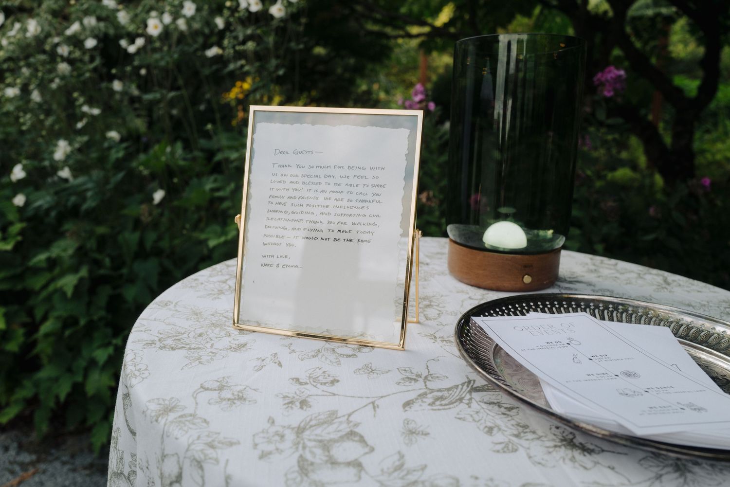 A framed text display on a white tablecloth with garden elements in the background.